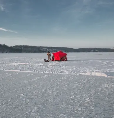 Fishing 3 tent campingAn ice fishing tent set up in the middle of a snowy lake, with a person fishing outside.